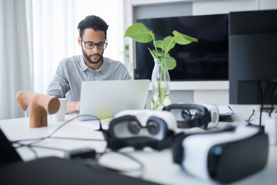 Man focused on laptop with VR goggles on desk, modern office setting.